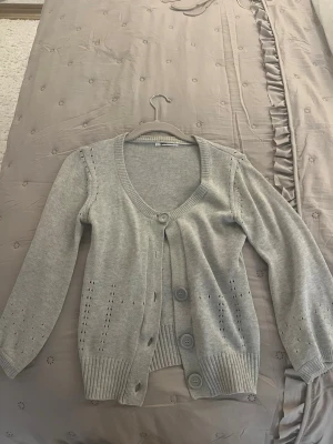 Ljusgrå kofta med stora knappar - Ljusgrå stickad kofta med v-ringning och tre stora knappar framtill. Koftan har ribbade muddar och nederkant samt dekorativa hålstickade detaljer längs ärmar och sidor. Perfekt för lager på lager och enkel att matcha.