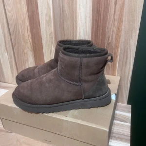 Bruna UGG boots i mocka - Mysiga bruna boots från UGG i mjuk mocka med kort skaft och rund tå. Insidan är fodrad med fluffigt material för extra värme och komfort. Klassisk UGG-design med grov sula och dragflik bak. Nypris 2299
