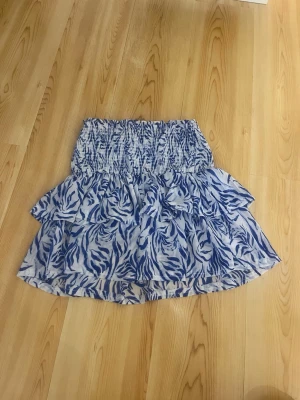 Blåvit volangkjol - Supersöt kort kjol med blått och vitt mönster. Kjolen har bred smock i midjan och dubbla volanger som ger en luftig och lekfull känsla. Perfekt för varma dagar och enkel att matcha med en enkel topp.