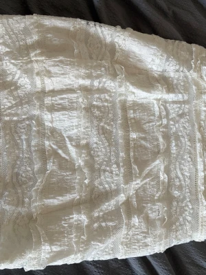 Vit spetskjol med volangdetaljer - Säljer en romantisk vit kjol med spets och volangdetaljer. Kjolen har ett vackert mönster av spets i vertikala ränder och är perfekt för dig som gillar en feminin och bohemisk stil. Passar till både sneakers och sandaler. Köpt second hand så inte använd av mig! Storlek S
