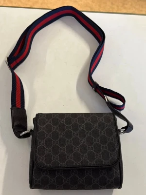 Svart axelväska från Gucci med GG-mönster - Snygg svart axelväska från Gucci med klassiskt GG-mönster över hela väskan. Väskan har en bred, justerbar axelrem i rött och blått tyg och är tillverkad i canvasmaterial. Perfekt för dig som vill ha en stilren och trendig accessoar med lyxig känsla.