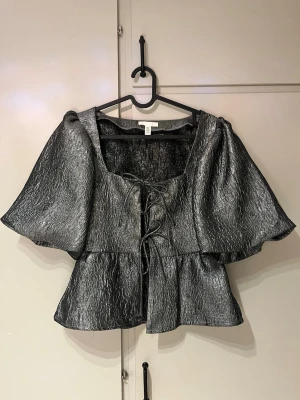 Silvrig blus med puffärm och snörning - Silvrig blus med markerad struktur, puffiga korta ärmar och snygg snörning framtill. Modellen är croppad och har en lätt glansig finish som ger en cool look. 