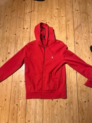 Röd hoodie från Polo Ralph Lauren - Snygg röd hoodie från Polo Ralph Lauren med dragkedja framtill och klassisk huva. Tröjan har två fickor på magen och den ikoniska lilla loggan broderad på bröstet. Tillverkad i mjukt bomullsmaterial som är skönt att ha på sig.