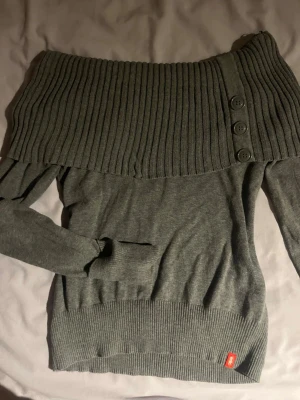 Grå off-shoulder stickad tröja - Snygg grå stickad tröja med bred ribbad off-shoulder-krage och tre dekorativa knappar. Tröjan har långa ärmar och ribbade muddar vid ärmslut och nederkant. Liten röd detalj nertill. Perfekt för en trendig look.