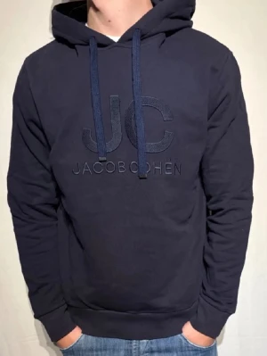 Mörkblå hoodie från Jacob Cohën - Snygg mörkblå hoodie från Jacob Cohën med stor huva och dragsko. Framtill finns ett diskret broderat JC-logotyp och märkesnamnet. Hoodien har ribbade muddar vid ärmar och nederkant. Perfekt för en avslappnad och trendig stil.