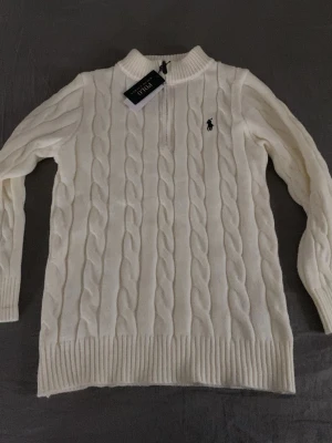 Vit kabelstickad tröja Polo Ralph Lauren - Kabelstickad vit tröja från Polo Ralph Lauren med halv dragkedja och ribbad krage. Tröjan har det klassiska broderade logotypen på bröstet och är gjord i mjukt material. Perfekt för dig som gillar stilrena och tidlösa plagg. Oskadad i perfekt skick använt 1 gång storlek M passar både S och M