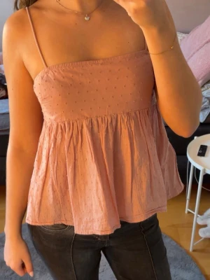 Rosa linnetopp med tunna band - Söt ljusrosa linnetopp med tunna axelband och lätt volang nedtill. Toppen har små diskreta prickar i tyget och är luftig med en flowy passform. Perfekt för varma dagar och enkel att styla med jeans eller kjol.