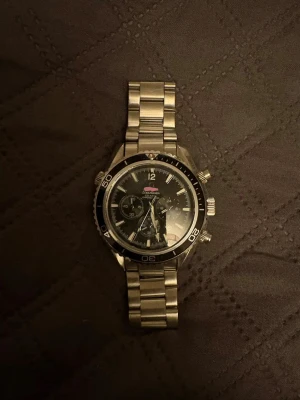 Silverfärgad kronografklocka Omega - Stilren silverfärgad kronografklocka från Omega med svart urtavla och tydliga markeringar. Klockan har robust länkarmband i metall och flera funktionella urtavlor. Perfekt accessoar för dig som gillar klassisk och sportig stil.