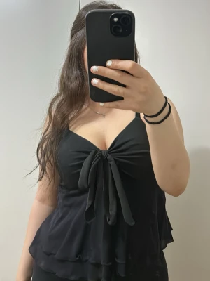 Svart linnetopp med knyt fram - Säljer en svart linnetopp med smala axelband och en stor knyt-detalj framtill. Toppen har volangkant nedtill och en lätt v-ringad urringning. Den är inte använd, endast testad💗