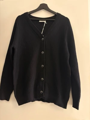 Svart V-ringad cardigan XL - Svart cardigan i storlek XL. Tröjan har V-ringning, knappar framtill och en diskret stickad struktur. Perfekt för lager-på-lager och enkel att matcha med olika outfits. Oanvänd.