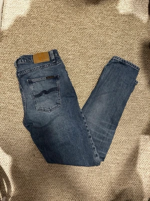 Blå raka jeans från Nudie Jeans - Säljer ett par klassiska blå jeans från Nudie Jeans med rak passform och fem fickor. Jeansen har en snygg tvättad look, kontrastsömmar och Nudie-loggan på bakfickan. Tillverkade i slitstark denim av bomull.