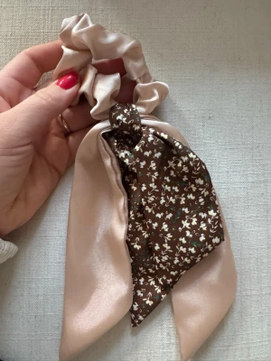 Beige scrunchie med brun scarfdetalj - Snygg scrunchie i glansigt beige tyg med fastsydd scarfdetalj. Scarfen är brun med små vita blommor och ger en trendig look. Perfekt för att piffa till din hästsvans eller knut. Materialet känns mjukt och lyxigt.
