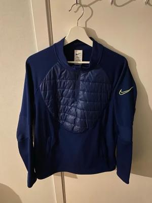 Mörkblått Nike träningsset - Snyggt mörkblått träningsset från Nike med matchande tröja och byxor. Tröjan har quiltad front, halv dragkedja och gul Nike-logga på ärmen. Byxorna har resår i midjan och gul Nike-logga på benet. Perfekt för träning eller chill.