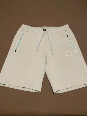 Ljusgrå shorts från BOSS - Snygga ljusgrå shorts från BOSS med elastisk midja och dragsko. Två fickor med ljusblå dragkedjor och diskret BOSS-logga i vitt på ena benet. Perfekta för en avslappnad och trendig stil. Tillverkade i mjukt bomullsmaterial.