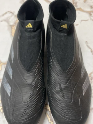 Adidas Predator svarta fotbollsskor - Säljer ett par svarta Adidas Predator fotbollsskor med klassiska tre ränder i silver på sidan. Skorna har strumpliknande skaft och är tillverkade i syntetmaterial för bra passform och kontroll. Perfekta för dig som vill ha stilrena och moderna skor på planen. De känns lite mindre i storlek, jätte fina och knappt använda. Perfekt skor som inte blir smutsiga 