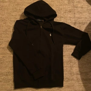 Svart hoodie från Polo Ralph Lauren - Svart hoodie med dragkedja från Polo Ralph Lauren. Klassisk design med huva och dragsnören, samt den ikoniska broderade loggan på bröstet. Mjuk bomullskänsla och fickor framtill. Perfekt för en avslappnad och stilren look.