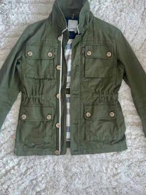 Field jacket/wind breaker J Lindeberg - Grön field jacket/windbreaker från J Lindeberg. Jackan är relativt tunn och är i allmänt bra skick. Har ett litet ställe med lite ställningar men enligt mig gör det jackan snyggare. 