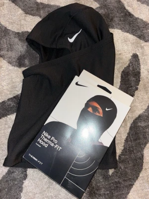 Nike Pro Therma-FIT balaclava svart - Nike Pro Therma-FIT balaclava i svart funktionsmaterial. Huva med tight passform och vit Nike-logga framtill. Perfekt för träning utomhus eller kalla dagar. Tillverkad i stretchigt, värmande material som andas.