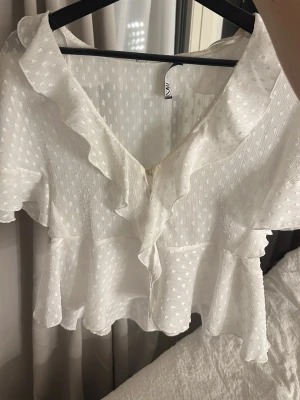 Vit volangblus från NA-KD - Söt vit blus från Zara med volangdetaljer längs ringningen och prickigt strukturmönster i tyget. Blusen har korta ärmar och lätt v-ringad hals. Tillverkad i ett tunt, luftigt material som ger en romantisk känsla.