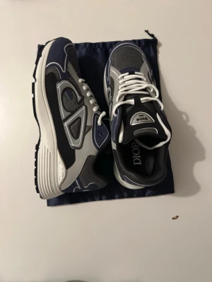 Dior B30 sneakers i blå och grå - Snygga Dior B30 sneakers i blått, grått och svart med vita detaljer. Skorna har en chunky sula, meshpaneler och stora Dior-loggor på sidan och tungan. Perfekt för dig som gillar exklusiva och trendiga sneakers med sportig vibe.
