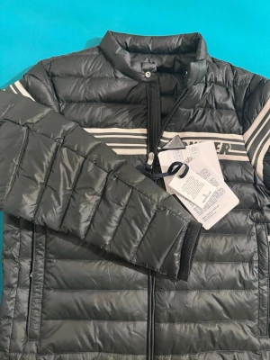 Grön dunjacka från Moncler - Moncler Renald med vita ränder och logga över bröstet. Jackan har dragkedja framtill, ståkrage och två sidofickor. Tillverkad i glansigt material och fylld med dun för extra värme. Perfekt för kalla vinterdagar. Endast testad men för liten på mig. NFC scan finns också, kvitto kan jag nog få fram.