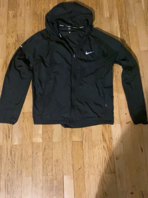 Svart vindjacka med huva från Nike - Snygg svart vindjacka från Nike med huva och dragkedja framtill. Jackan har en diskret Nike-logga på bröstet och är tillverkad i lätt, vindtätt material. Perfekt för löpning eller när du vill ha en sportig look.
