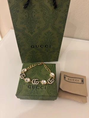 Gucci armband med pärlor och GG-logo - Lyxigt armband från Gucci med guldfärgad kedja, stora glittrande pärlor och ikoniska GG-loggor dekorerade med stenar. Armbandet har en exklusiv och trendig design som verkligen sticker ut. Perfekt accessoar för dig som gillar statement-smycken.