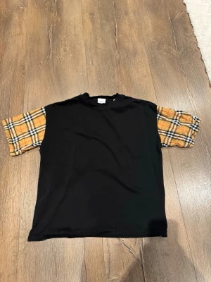 Svart Burberry t-shirt med rutiga ärmar - Säljer en svart t-shirt från Burberry med klassiska beige-rutiga kortärmar. T-shirten har en rak passform och är tillverkad i mjuk bomull. Perfekt för dig som vill ha en unik och stilren look med en touch av Burberrys ikoniska mönster.