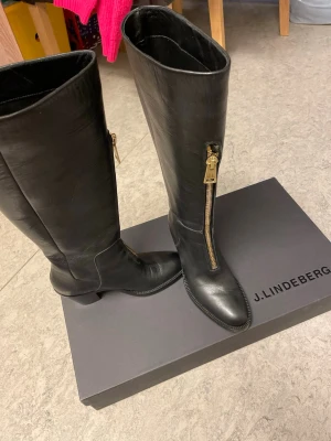 Svarta läderstövlar från J.Lindeberg - Snygga svarta knähöga stövlar från J.Lindeberg i äkta läder. Stövlarna har en markerad guldfärgad dragkedja på sidan och en låg klack. Tillverkade i Italien och har en stilren, klassisk design som passar till många outfits.
