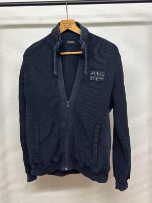 Mörkblå ziptröja från Napapijri - Mörkblå ribbstickad ziptröja från Napapijri med hög krage och dragsko. Tröjan har två fickor framtill och en broderad flagga på bröstet. Perfekt för dig som gillar en sportig och avslappnad stil.