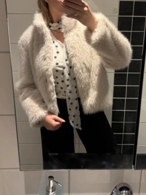 Beige fluffig pälsjacka - Supermjuk och fluffig beige pälsjacka med rak passform och långa ärmar. Jackan har en klassisk krage och stängs framtill. Perfekt för att hålla värmen under kalla dagar och ger en trendig look till din outfit.