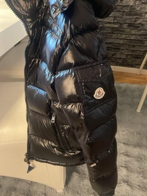 Svart glansig dunjacka från Moncler - Snygg svart dunjacka från Moncler med glansig finish, stor ficka med logotyp på ärmen och dragkedja framtill. Jackan har en klassisk puffer-design och är perfekt för kalla vinterdagar. Materialet är typiskt för Monclers exklusiva dunjackor.