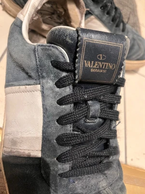 Valentino Garavani blåa sneakers - Snygga sneakers från Valentino Garavani i blå mocka med vita läderdetaljer och svart snörning. Klassisk låg modell med beige sula och diskret logga på plösen. Perfekta för dig som gillar exklusiva streetwear-vibbar.