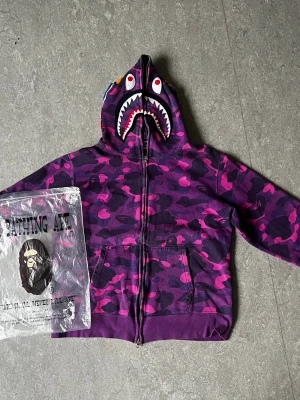 Purple BAPE Zip-Up hoodie - Säljer en ikonisk BAPE hoodie i lila färg! Dragkedjan går hela vägen över huvan.  broderade bokstäver, tiger detalj och hajdetaljer på huvan! storlek XL men sitter som large! Kommer med dustbag