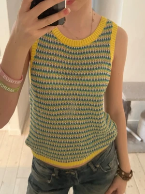 Gul och blå stickad linnetopp - Storlek M men passar mig som har XS och S!! Säljer en färgglad stickad linnetopp med gul kant och ett mönster i blått, grönt och vitt. Toppen är ärmlös och har en rund halsringning. Perfekt för dig som vill ha en unik och retroinspirerad look.