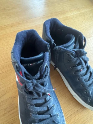 Marinblå Tommy Hilfiger sneakers - Snygga marinblå sneakers från Tommy Hilfiger med högt skaft och klassisk logga i rött, vitt och blått på sidan. Skorna har vita sulor med mönster och är gjorda i ett slitstarkt material med vadderad insida för extra komfort.