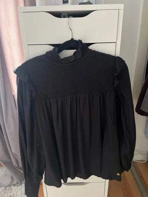 Svart volangblus med hög krage - Snygg svart blus med smock upptill, volangdetaljer vid axlarna och hög krage. Långa ärmar med breda muddar och lös passform nedtill. Perfekt för dig som gillar en trendig och stilren look. Från Pernilla Wahlgren 