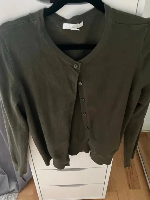 Olivgrön kofta med rund hals - En stilren olivgrön kofta med rund halsringning och knäppning framtill. Långa ärmar och ribbade muddar vid ärmslut och nederkant. Perfekt att slänga över en t-shirt eller linne för en avslappnad look.