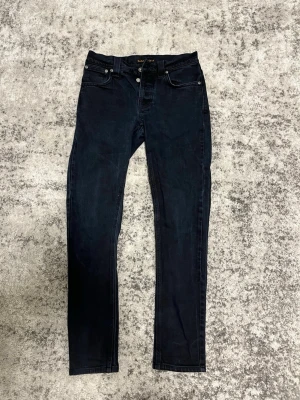 Mörkblå straight jeans från Nudie - Säljer ett par mörkblå straight jeans från Nudie Jeans med klassisk femficksmodell och normal passform. Jeansen har svarta sömmar och knappgylf. Perfekta för en stilren och avslappnad look. W28 L32