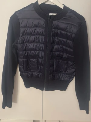 Mörkblå quiltad cardigan med dragkedja - Snygg mörkblå cardiganjacka med quiltad framsida och stickade ärmar samt rygg. Jackan har två framfickor, ribbade muddar och dragkedja framtill. Perfekt för lager på lager och en cool vardagslook.