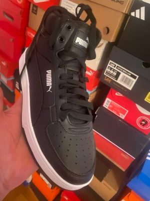 Svarta Puma high top sneakers - Stilrena svarta high top sneakers från Puma med vit sula och klassisk logga på sidan och plösen. Skorna har perforerad tå, svarta skosnören och är tillverkade i syntetmaterial och läderimitation. Perfekta för dig som gillar sportig och clean stil.