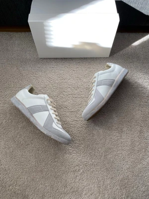 Maison Margiela White Paint Gats - Stilrena vita sneakers från Maison Margiela med beige och grå detaljer. Skorna har lågt skaft, klassisk snörning och ovandel i läder och mocka. Sula i gummi för bra grepp. Perfekta för dig som gillar minimalistisk och clean stil.