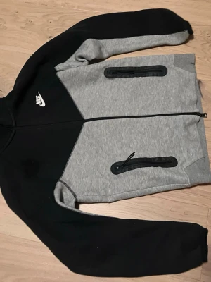 Nike svart och grå hoodie - Snygg hoodie från Nike i svart och grått med dragkedja framtill och två vertikala fickor med dragkedja. Klassisk Nike-logga på bröstet och huva för extra stilpoäng. Perfekt för dig som gillar sportig och avslappnad look.