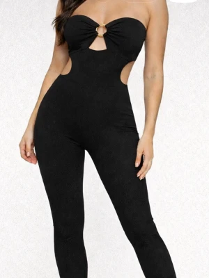 Svart strapless jumpsuit med cut-outs - Säljer en svart jumpsuit med strapless design och snygga cut-outs vid midjan. Framtill finns en ringdetalj som ger en cool look. Tight passform och långa ben, perfekt för dig som vill sticka ut. Materialet är stretchigt och bekvämt.