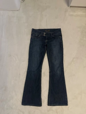 Mörkblå bootcut low waist jeans True2U - Snygga mörkblå bootcut jeans från True2U med dubbla silverknappar i midjan och klassiska fem fickor. Jeansen har en lätt utsvängd passform med låg midja och är tillverkade i denim med kontrastsömmar. Perfekta för en trendig och avslappnad look.