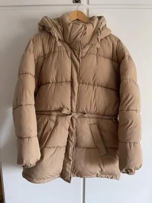 Beige pufferjacka från Bik Bok - Beige pufferjacka från Bik Bok med huva och dragsko i midjan. Jackan har två stora fickor framtill och stängs med dragkedja och tryckknappar. Perfekt för kalla vinterdagar tack vare den tjocka vadderingen och den långa modellen.