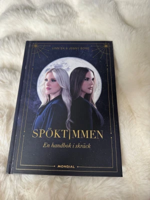Spöktimmen: En handbok i skräck - Säljer boken 'Spöktimmen: En handbok i skräck' av Linn Ek och Jenny Borg. Boken har hård pärm i mörkblå färg med guldtryck och ett mystiskt omslag med måne och två personer. Perfekt för dig som gillar skräck, mysterier och true crime.