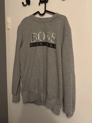 Grå sweatshirt från Hugo Boss - Säljer en grå sweatshirt från Hugo Boss med stort BOSS-tryck på bröstet och klassisk rund halsringning. Tröjan har långa ärmar och ribbade muddar vid ärmslut och nederkant. Perfekt för en chill och avslappnad look.