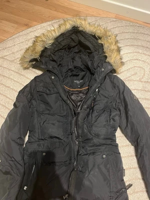 Svart parkas från Hollies med fuskpäls - Snygg svart parkas från Hollies med stor huva och fuskpäls runt kanten. Jackan har flera extra fickor, dragkedja och knappar framtill samt justerbar midja. Perfekt för kalla dagar och riktigt trendig look. Pris kan diskutera.
