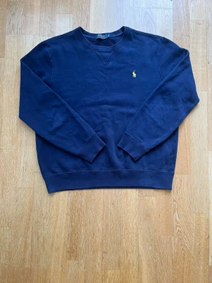 Mörkblå sweatshirt från Polo Ralph Lauren - Klassisk mörkblå sweatshirt från Polo Ralph Lauren med rund halsringning och gul broderad logga på bröstet. Tröjan har ribbade muddar vid ärmslut och nederkant. Perfekt för en avslappnad och stilren look.